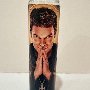 May include: A clear glass prayer candle featuring a printed image of a man with short dark hair, wearing a dark garment and a silver cross necklace. The man is shown with his hands pressed together in a prayer position against a golden halo background.