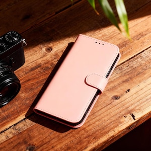 May include: A light pink phone case with a magnetic closure and a stitched border. The case is made of a smooth material and has a curved tab for securing the case closed. A black camera is visible in the background.