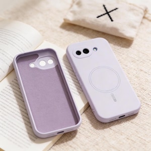 Può includere: Due custodie per telefono lilla, una aperta e una chiusa, sono esposte su un libro. La custodia aperta rivela un interno viola chiaro. La custodia chiusa presenta un design circolare sul retro e un ritaglio per la fotocamera.