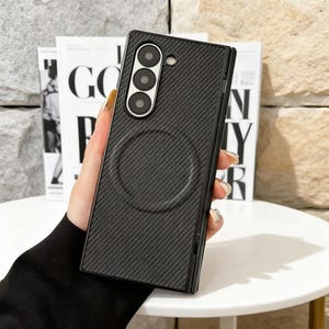 May include: Black phone case with a carbon fiber texture and a circular indentation. The case has a triple-lens camera cutout and a sleek, modern design. The phone case is held in a person's hand.