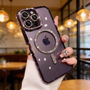 May include: A purple phone case with a clear back and a decorative border of sparkling rhinestones. The case features a ring of rhinestones around the camera and a magnetic circle with the Apple logo, also adorned with rhinestones.