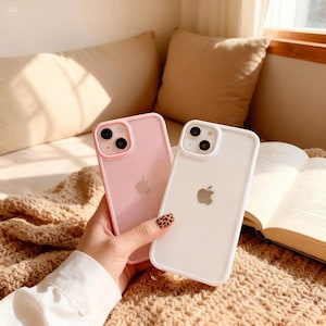 May include: Two iPhone cases, one pink and one white, are displayed. The pink case has a pink border, and the white case has a white border. Both cases feature the Apple logo. The background is neutral.