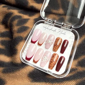 Könnte beinhalten: Ein Set künstlicher Fingernägel in einer transparenten Kunststoffbox. Die Nägel weisen verschiedene Designs auf, darunter eine French-Spitze mit dunkelrotem Rand, eine hellrosa Basis mit Leopardenmuster und eine hellrosa Basis mit Perlen. Der Text "Handmade Nails" ist sichtbar.