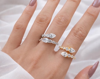 May include: Two rings, one silver and one gold, each adorned with clear, pear-shaped and round gemstones. The silver ring wraps around the finger, while the gold ring has a similar design. The rings are displayed on a hand with manicured nails.