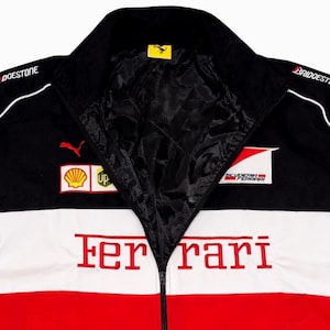 May include: Black, white, and red Ferrari racing jacket with a zip-up front. The jacket features sponsor logos, including Puma and Shell, and the Ferrari logo. The jacket has a black collar and lining.
