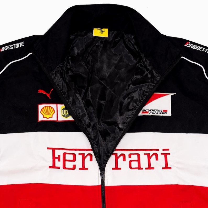 May include: Black, white, and red Ferrari racing jacket with a zip-up front. The jacket features sponsor logos, including Puma and Shell, and the Ferrari logo. The jacket has a black collar and lining.