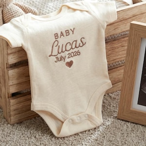 May include: A cream-coloured baby bodysuit featuring brown embroidery that reads "BABY Lucas July 2026" with a small heart icon. The bodysuit is placed in a wooden crate with a crocheted bunny, a beige pacifier, and a framed ultrasound image.