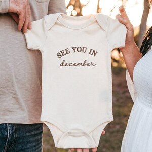 Natural baby bodysuit held by parents outdoors with “SEE YOU IN december” embroidery.
Dark brown thread lettering centered on chest, soft neutral fabric with pregnancy announcement vibe.