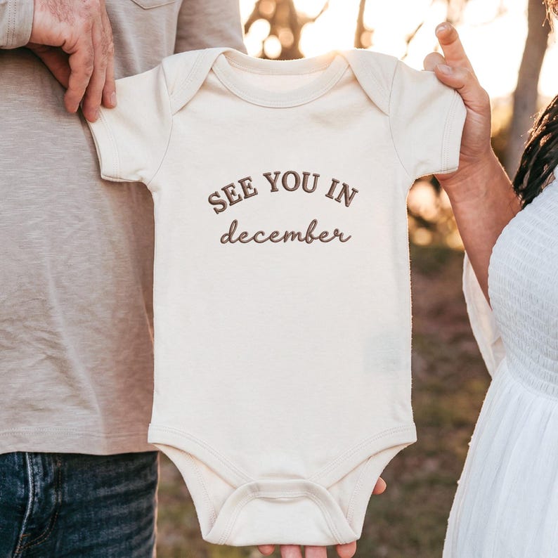 Natural baby bodysuit held by parents outdoors with “SEE YOU IN december” embroidery.
Dark brown thread lettering centered on chest, soft neutral fabric with pregnancy announcement vibe.