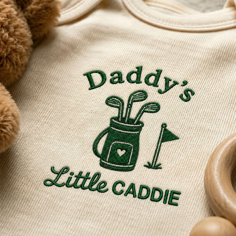 May include: Cream-colored baby onesie with green embroidered text and a golf-themed design. The text reads "DADDY'S LITTLE CADDIE" above a graphic of golf clubs in a golf bag. A wooden toy is visible in the corner.