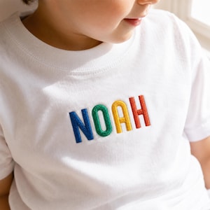 May include: A white short-sleeved t-shirt featuring the name NOAH embroidered across the chest in block letters. Each letter is stitched in a different vibrant colour, with N in blue, O in green, A in yellow, and H in orange thread.