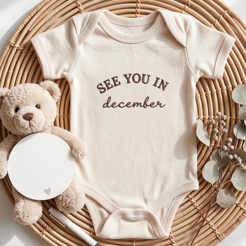 Natural baby onesie with “SEE YOU IN december” embroidery on middle chest.
Dark brown thread text in a mix of uppercase and script font, styled flatlay with neutral baby props.