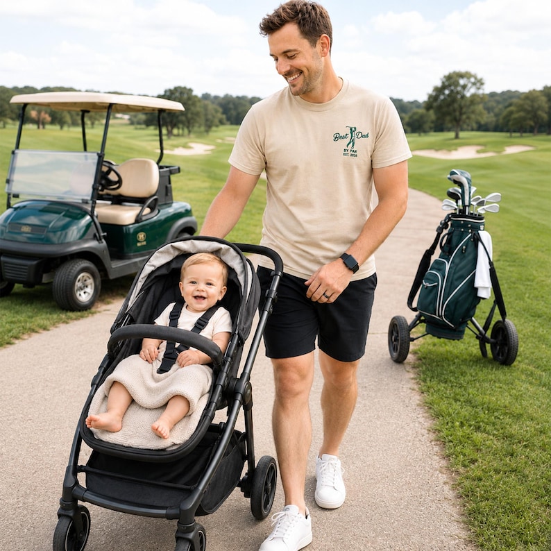 May include: A father and baby wearing matching beige golf-themed shirts. The father's shirt says "Best Dad by Par Est. 2026." The baby's onesie says "Daddy's Little Caddie." A golf ball and golf bag are in the background.