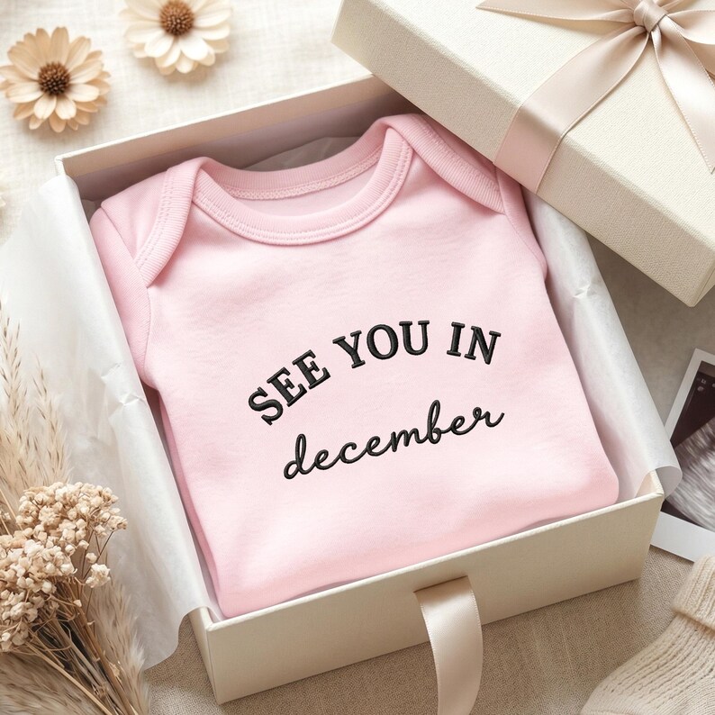 Pink baby onesie in gift box with “See You In December” embroidery in black thread.
Centered chest design with minimalist font, styled as baby shower or pregnancy announcement gift.