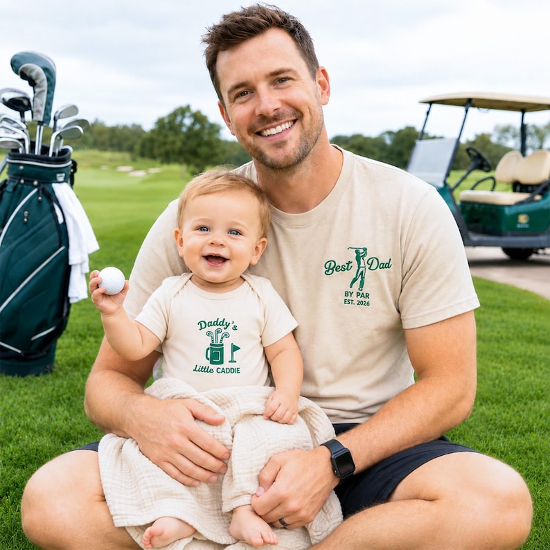 May include: A father and baby wearing matching beige golf-themed shirts. The father's shirt says "Best Dad by Par Est. 2026." The baby's onesie says "Daddy's Little Caddie." A golf ball and golf bag are in the background.