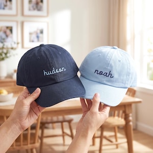 May include: Two baseball caps made of cotton fabric. The left cap is dark navy blue with the name "hudson" embroidered in light blue thread. The right cap is light blue with the name "noah" embroidered in dark navy blue thread.