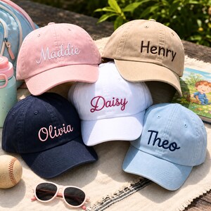 May include: Five baseball caps in pink, tan, white, navy blue, and light blue. Each cap features embroidered names: Maddie in white, Henry in dark brown, Daisy in magenta, Olivia in light pink, and Theo in dark blue, all in script or print.
