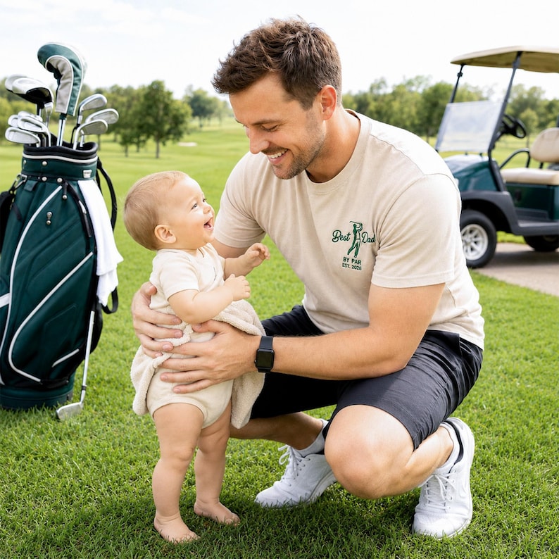 May include: A father and baby wearing matching beige golf-themed shirts. The father's shirt says "Best Dad by Par Est. 2026." The baby's onesie says "Daddy's Little Caddie." A golf ball and golf bag are in the background.