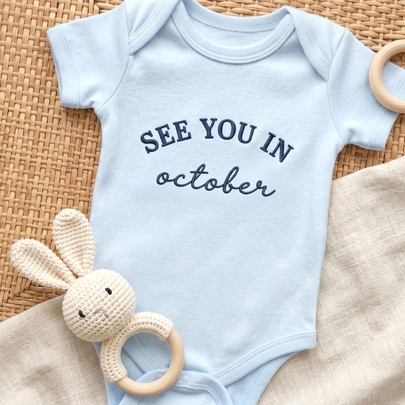 Light blue baby onesie featuring “SEE YOU IN october” embroidery on chest.
Navy thread lettering in mixed font styles, styled flatlay with soft neutral accessories.
