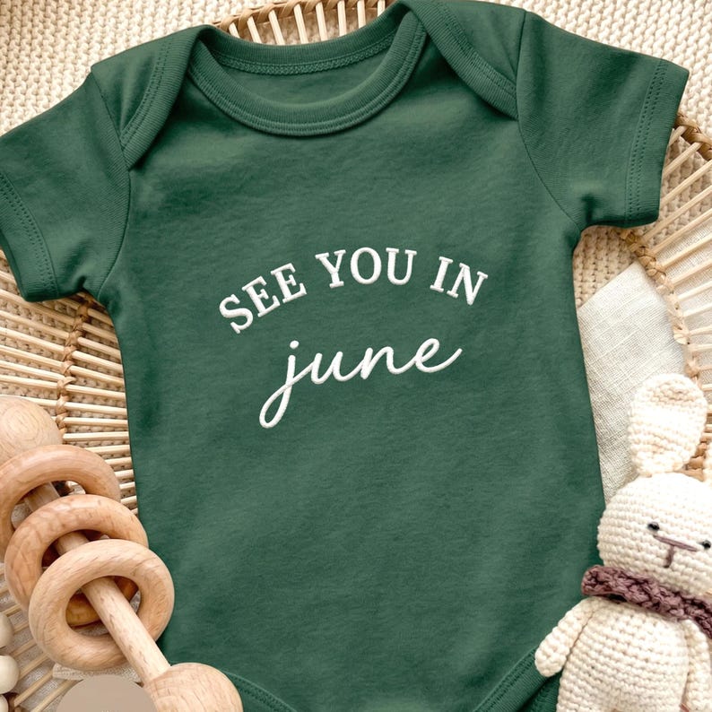 Forest green baby onesie with “SEE YOU IN june” embroidery on middle chest.
White thread text in clean uppercase and script font, styled with wooden toys and cozy baby decor.