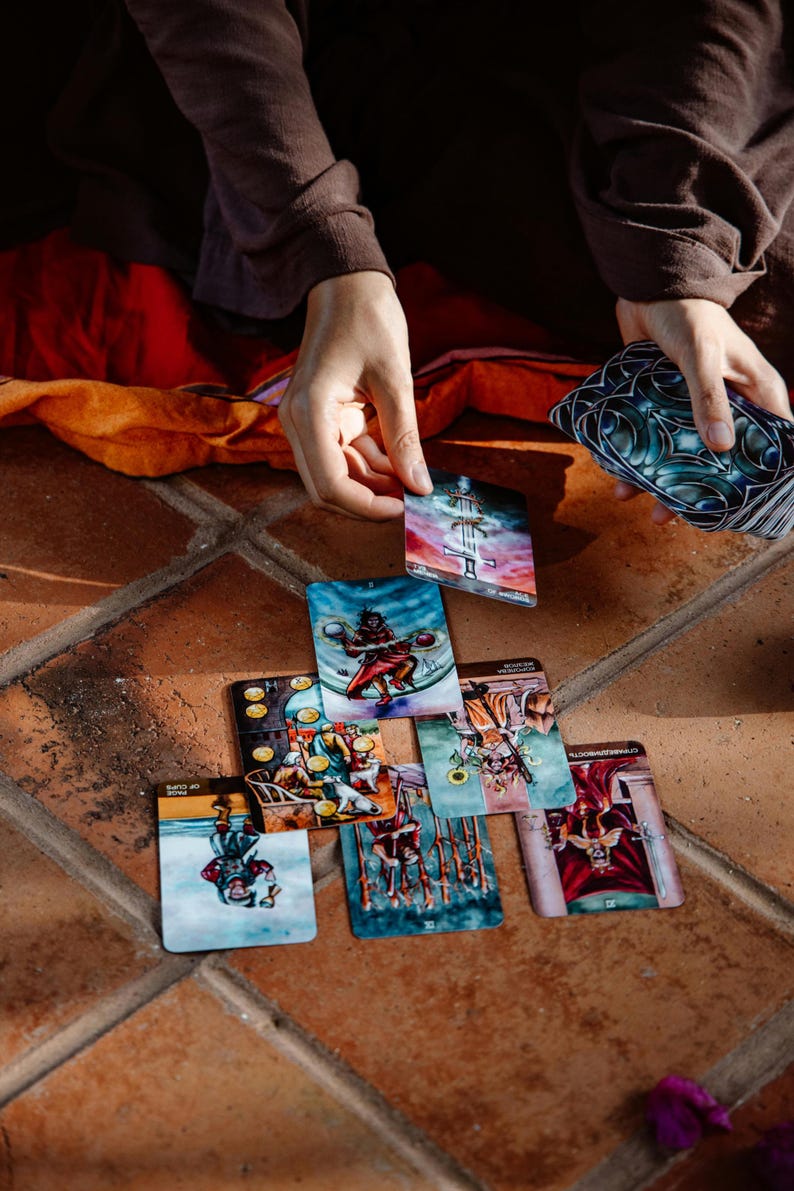 May include: A collection of tarot cards with colorful illustrations, including images of figures, symbols, and scenes. The cards are arranged on a terracotta tile surface, with hands holding and selecting cards from the deck. The cards feature various colors and designs.