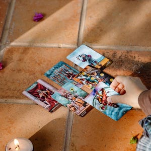 May include: A collection of tarot cards with colorful illustrations, including images of figures and symbols, are arranged on a terracotta tile surface. A lit candle and scattered flower petals are also present.