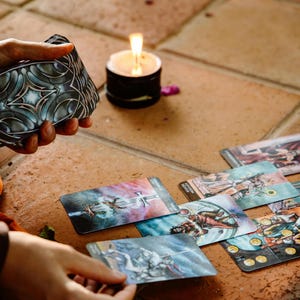May include: A deck of tarot cards with a decorative back design is held near several face-up cards. A lit candle sits nearby on a tiled surface. The cards feature colorful illustrations, suggesting a theme of divination and spirituality.