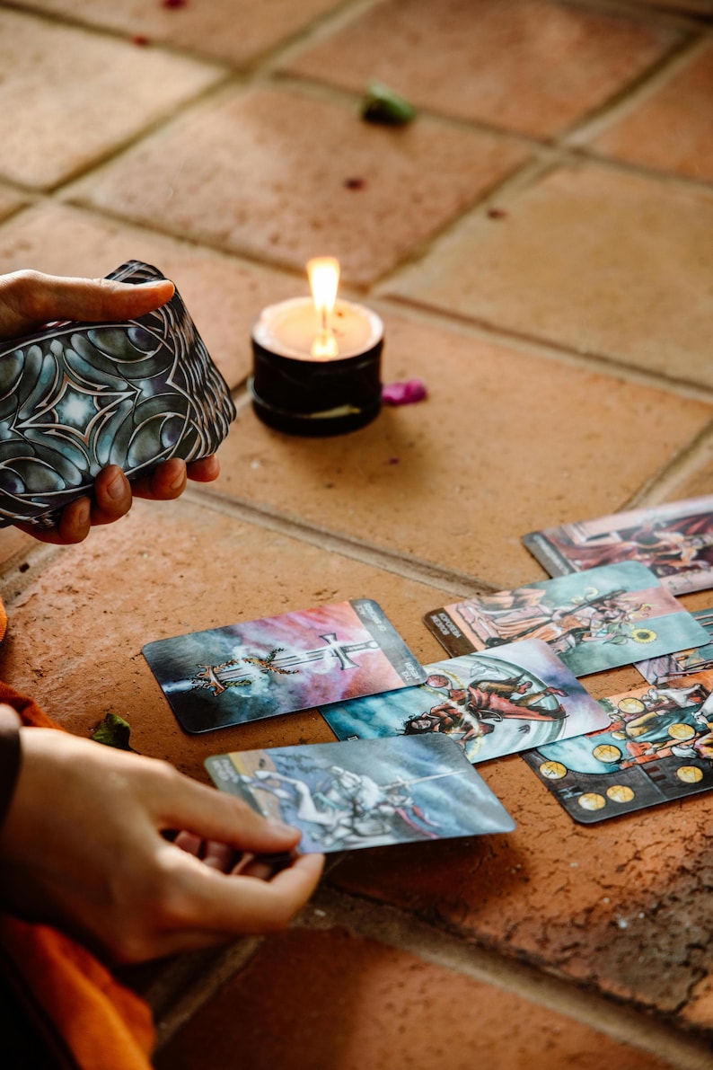 May include: A deck of tarot cards with a decorative back design is held near several face-up cards. A lit candle sits nearby on a tiled surface. The cards feature colorful illustrations, suggesting a theme of divination and spirituality.