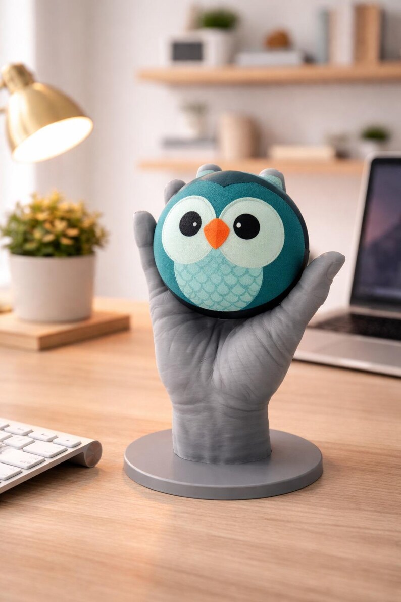 May include: A teal and blue owl-shaped stress ball with large white eyes and an orange beak. The owl is held by a gray hand-shaped stand on a wooden desk. A desk lamp, keyboard, and laptop are in the background.