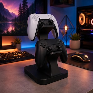 May include: A black and white gaming controller stand holding two controllers. The stand is black and has a curved design. One controller is white, the other is black. The stand is on a desk with a keyboard and mouse in the background.