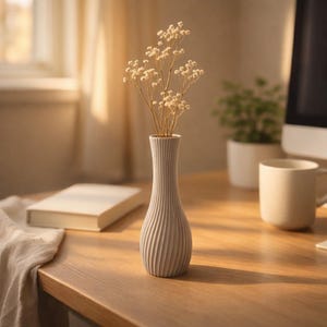 May include: A small, light gray vase with a ribbed design holds dried flowers. The vase is on a wooden table, with a book, a mug, and a small plant in the background. The vase is approximately 8 inches tall.
