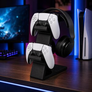 May include: A black gaming accessory stand holding two white game controllers and a black headset. The stand is on a wooden surface, with a gaming console and monitor in the background.