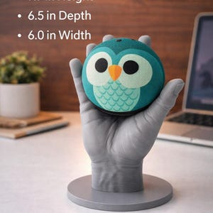 May include: A teal owl-shaped speaker with a white and black face and orange beak, held by a gray hand-shaped stand. The speaker is 7.9 inches high, 6.5 inches deep, and 6.0 inches wide. The speaker has a round shape and a fabric-like texture.