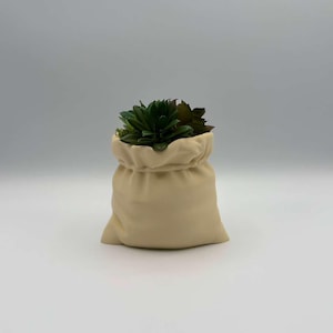 Puede incluir: Una maceta de cerámica beige con forma de bolsa con cordón, llena de suculentas artificiales verdes y marrones. La maceta tiene una superficie texturizada y está sobre un fondo blanco.