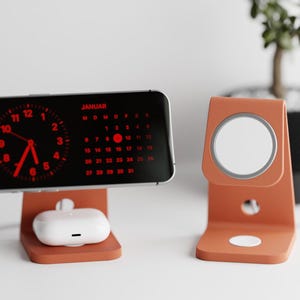May include: An orange charging stand with a phone displaying a red clock and calendar, and a white earbud case. A second orange stand holds a white charging pad. A small plant is in the background.