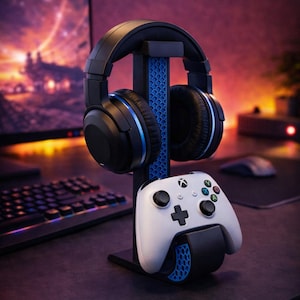 Puede incluir: Unos auriculares para juegos negros y azules y un mando Xbox blanco se muestran en un soporte negro y azul. El soporte está sobre un escritorio con un monitor de ordenador y un teclado en el fondo. Los auriculares tienen detalles azules.