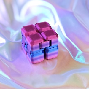May include: A colorful, gradient-colored infinity cube toy. The cube is made of multiple smaller cubes that are connected and can be manipulated. The colors transition from pink to blue. The background is a shiny, iridescent fabric.