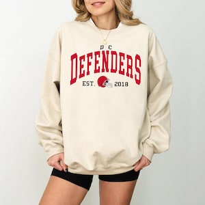 May include: A cream-colored sweatshirt with the text "DC DEFENDERS" in red, arched across the chest. Below the text is "EST. 2018" and a football helmet graphic. The sweatshirt has a crew neck and long sleeves.