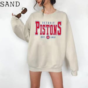 May include: A sand-colored sweatshirt featuring the text "DETROIT PISTONS" in red and blue lettering. The sweatshirt includes the team's logo and the year "1937". The long-sleeved top is a casual style.