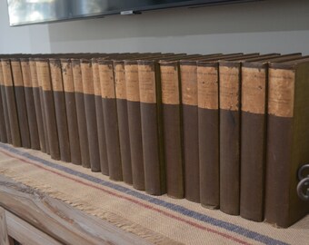 May include: A row of brown leather-bound books with gold lettering on the spines. The books are arranged on a shelf with a burlap runner.