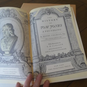 May include: Open book titled "The History of Tom Jones, A Foundling" by Henry Fielding. The book's cover features an illustration of a bust and ornate framing. Published by The Heritage Press, New York.