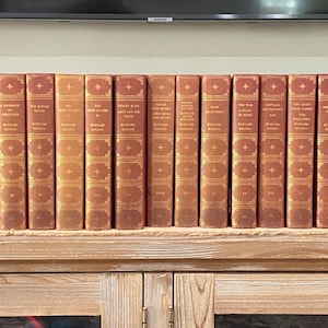 May include: A collection of 30 vintage hardback books with gold lettering on red spines. The books are arranged in a row on a wooden shelf.