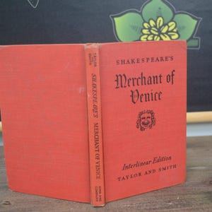 May include: A red hardcover book titled Shakespeare's Merchant of Venice, Interlinear Edition by Taylor and Smith. The front cover features black text and a small illustration of a face. The spine also displays the title and author information.