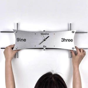 May include: A modern wall clock with a silver face and black hands. The clock face has the numbers 9ine, 12welve, 3hree, and 6ix. The clock is mounted on a white wall with horizontal and vertical metal bars.