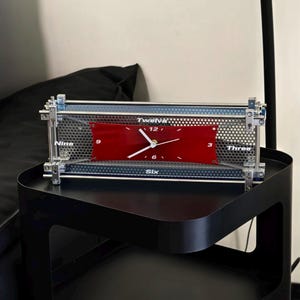 Peut inclure: Une horloge rectangulaire moderne avec un cadran rouge et des aiguilles et chiffres blancs. L'horloge est encadrée dans un cadre en métal argenté avec un écran en métal perforé. Le mot "Twelve" est imprimé en haut.
