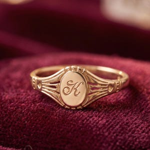 Op de afbeelding: Een goudkleurige ring met een ovale middenstuk met de letter "K" in een sierlijk handschrift. De ring heeft een gedetailleerd ontwerp met decoratieve elementen op de band en rond de centrale ovaal. De ring ligt op een dieprode fluwelen ondergrond.