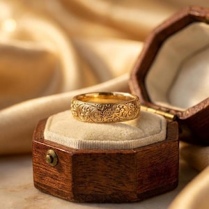Può includere: Un anello d'oro con un intricato motivo floreale è presentato in un portagioie ottagonale in legno aperto. L'anello è appoggiato su un cuscino color crema. Lo sfondo è un morbido tessuto dorato.