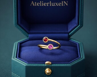 May include: A gold-colored ring with a red and pink gemstone design, presented in a teal jewelry box. The ring features a unique, open design with a red gemstone on one end and a pink gemstone on the other. The jewelry box has the text "AtelierluxeIN" on the inside.