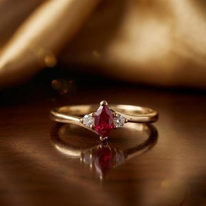 Op de afbeelding: Een gouden ring met een marquise-geslepen robijn als middensteen en twee kleinere diamanten aan weerszijden. De ring ligt op een reflecterend oppervlak met een gouden achtergrond.