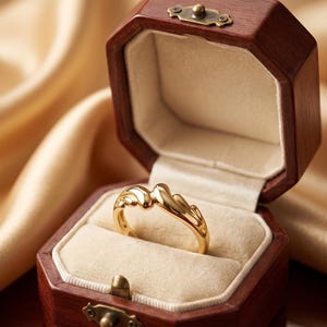 May include: A gold ring with a unique, flowing design rests inside an open, octagonal wooden jewelry box. The ring is the focal point, set against a cream-colored interior. The box is on a reflective, dark wooden surface.